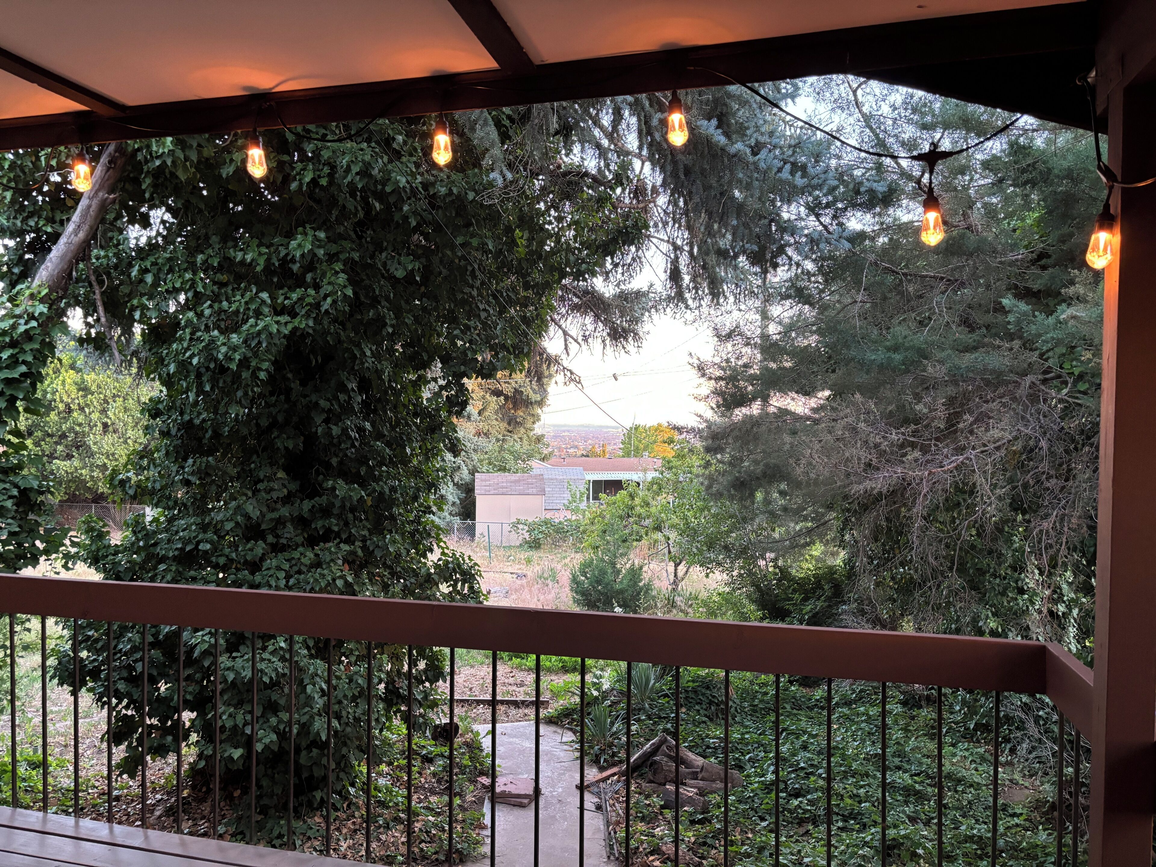 Un espacio al aire libre cubierto con barandilla de madera, luces colgantes y vistas a una casa y follaje circundante.