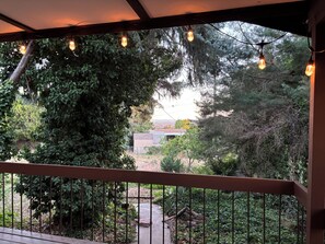 A covered outdoor area with a wooden railing, string lights, and a view of a house and surrounding greenery.