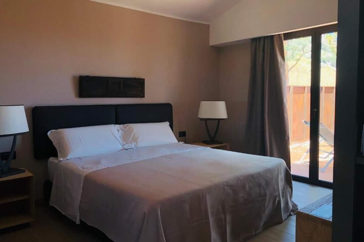 Hotelkamer met een groot bed, een nachtkastje met een lamp, een raam met gordijnen en uitzicht op het landschap.