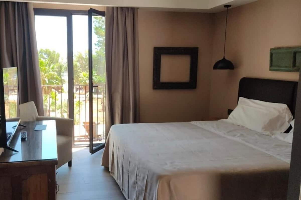 Een hotelkamer met een bed, een bureau, een stoel, een raam met gordijnen en uitzicht op een balkon.