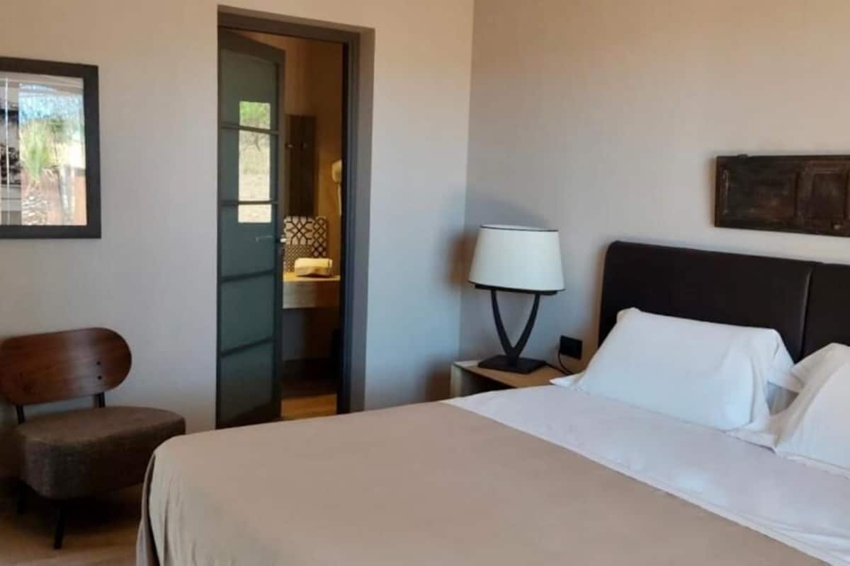 Een hotelkamer met een bed, een stoel, een nachtkastje met een lamp en een deur naar een andere kamer.