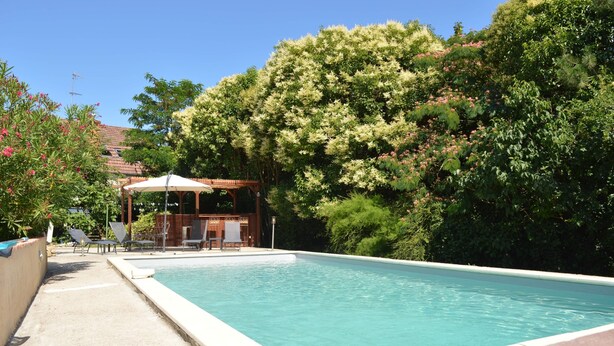 A rectangular swimming pool with a wooden deck and lounge chairs, surrounded by lush greenery and a gazebo.