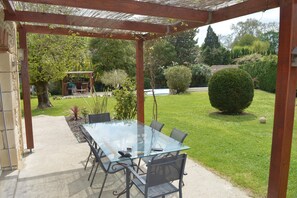 Ein Tisch mit Glasplatte und Stühlen auf einer Terrasses, umgeben von Grünpflanzen und einer Pergola.