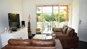 A living room with a brown leather sofa, a television, a small table, and a chair. There is a sliding glass door leading to an outdoor area with a table and chairs.