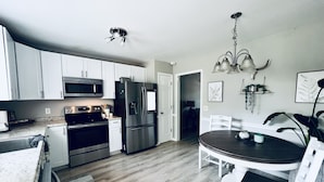 A modern kitchen with stainless steel appliances, white cabinetry, and a dining area with a wooden table and chairs. - The Nash Spot (Nashville)