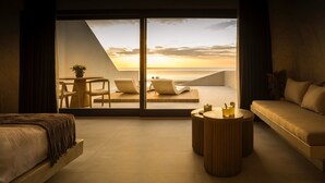 A modern hotel room with a large sliding glass door leading to a balcony with a sunset view. - Pnoé Luxury Suites (Santorini)