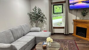 A living room with a grey sectional sofa, a round coffee table with a vase of flowers, a patterned rug, a fireplace, and a TV on the wall.