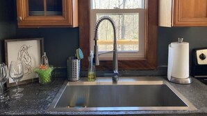 A kitchen with a stainless steel sink, wooden cabinets, a window with a view of trees, and various kitchen items on the counter.