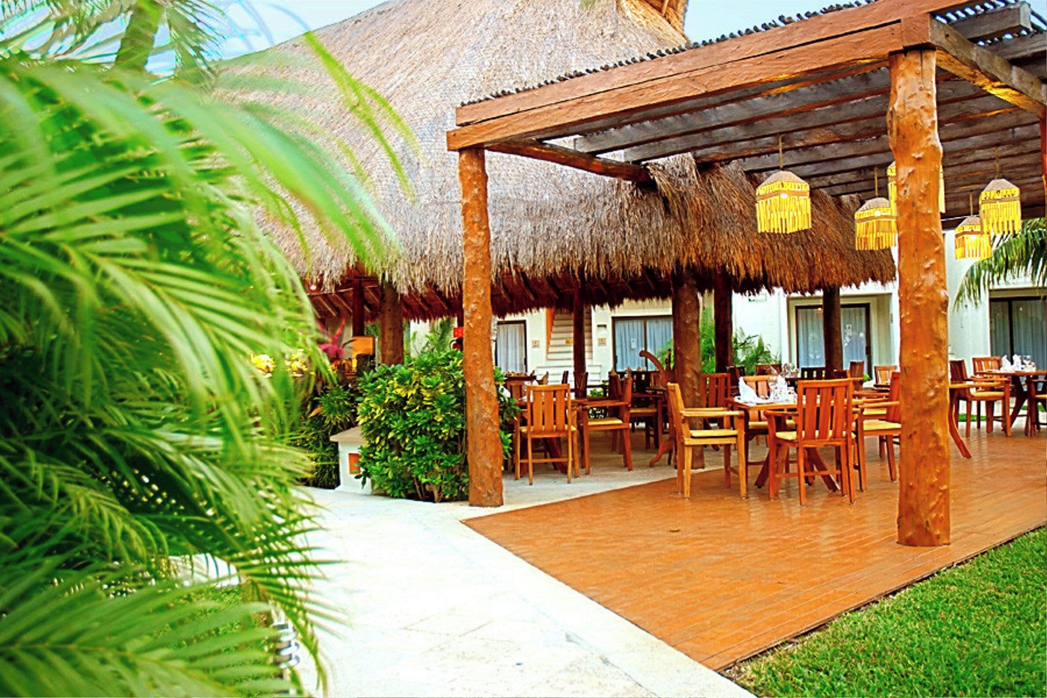 An outdoor dining area with wooden tables and chairs, a thatched roof structure, and tropical plants.
