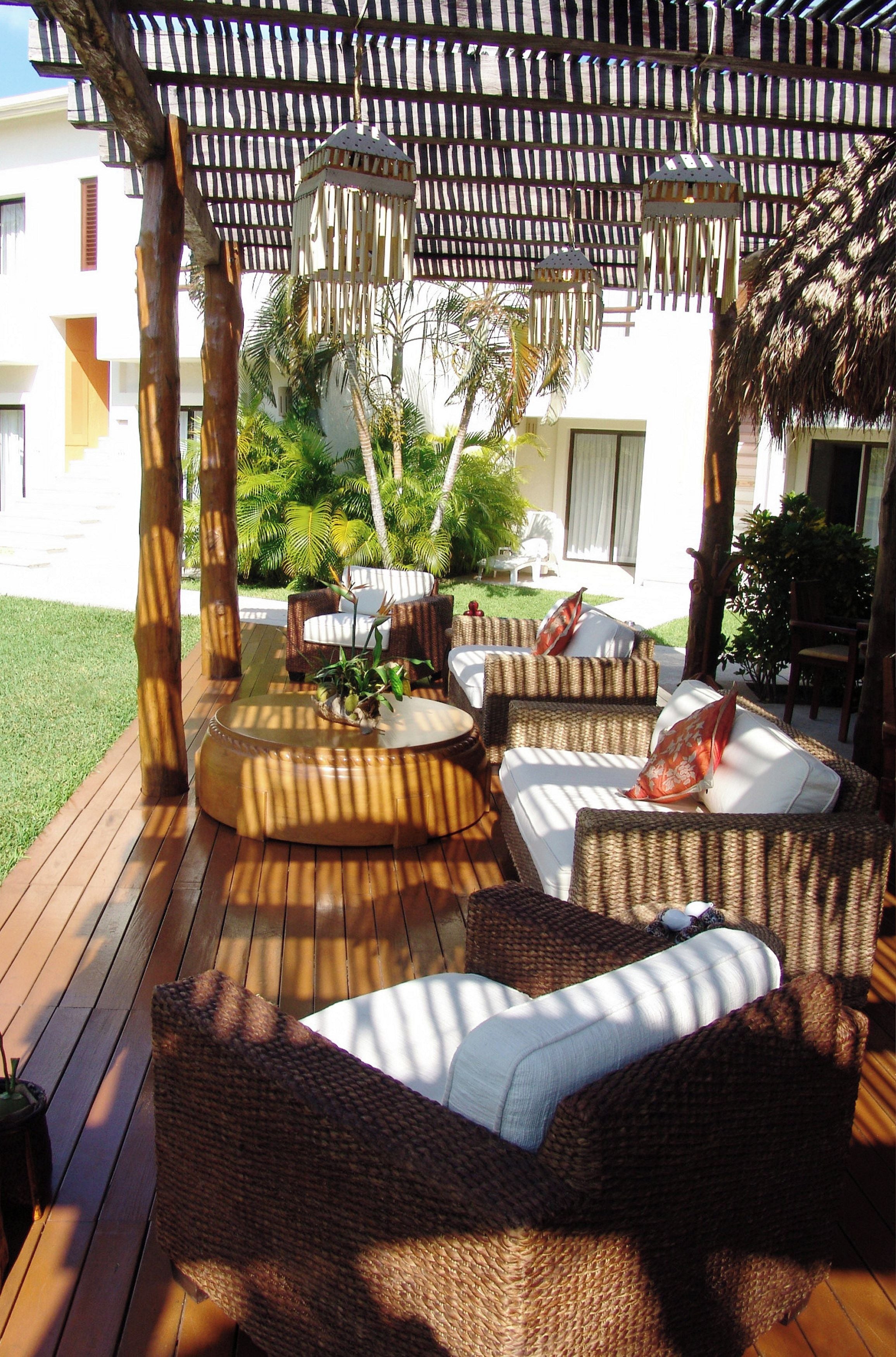 Un patio exterior con mobiliario de mimbre, una pérgola arriba y vista a un edificio blanco al fondo.
