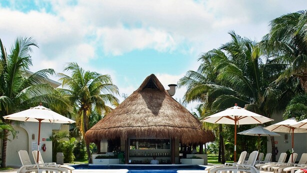 A swimming pool with lounge chairs and umbrellas, surrounded by palm trees and a thatched-roof structure. - Laguna Suite (Cancun)