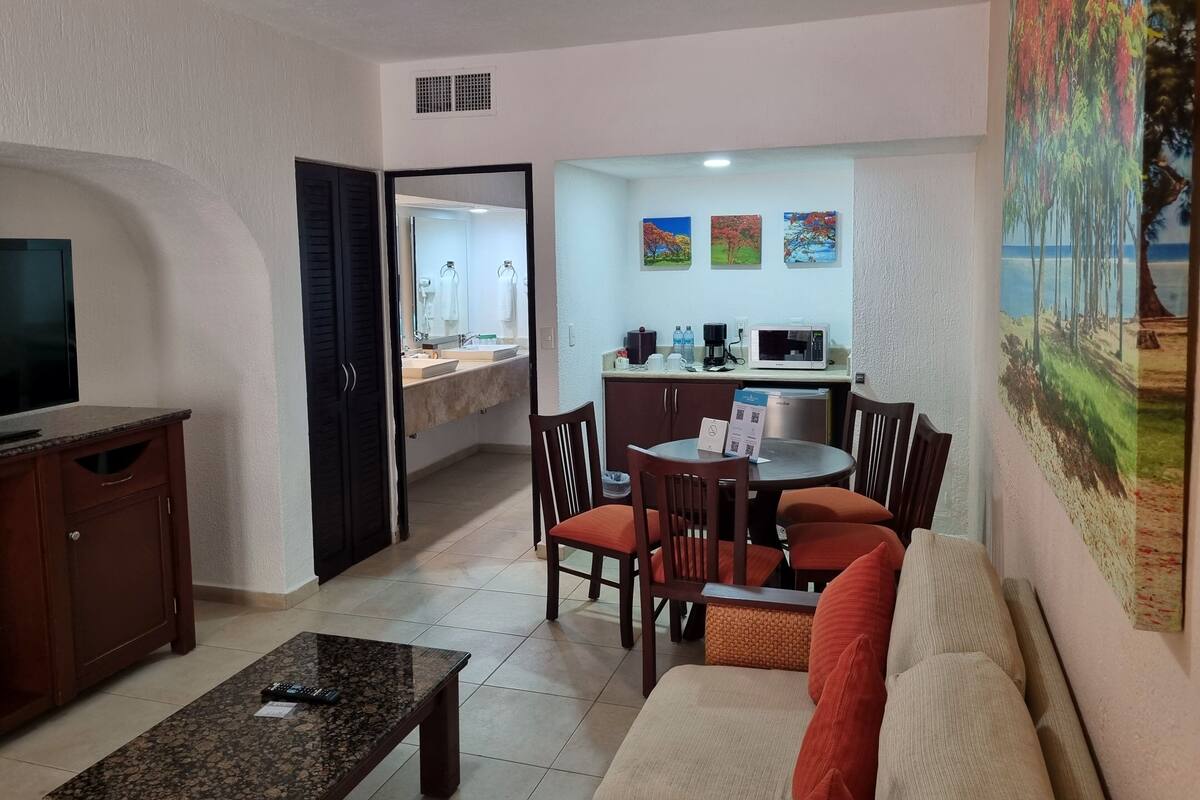 A living room with a TV, a sofa, and a coffee table. There is a dining area with a table and chairs. The kitchen is visible with a microwave and a sink.