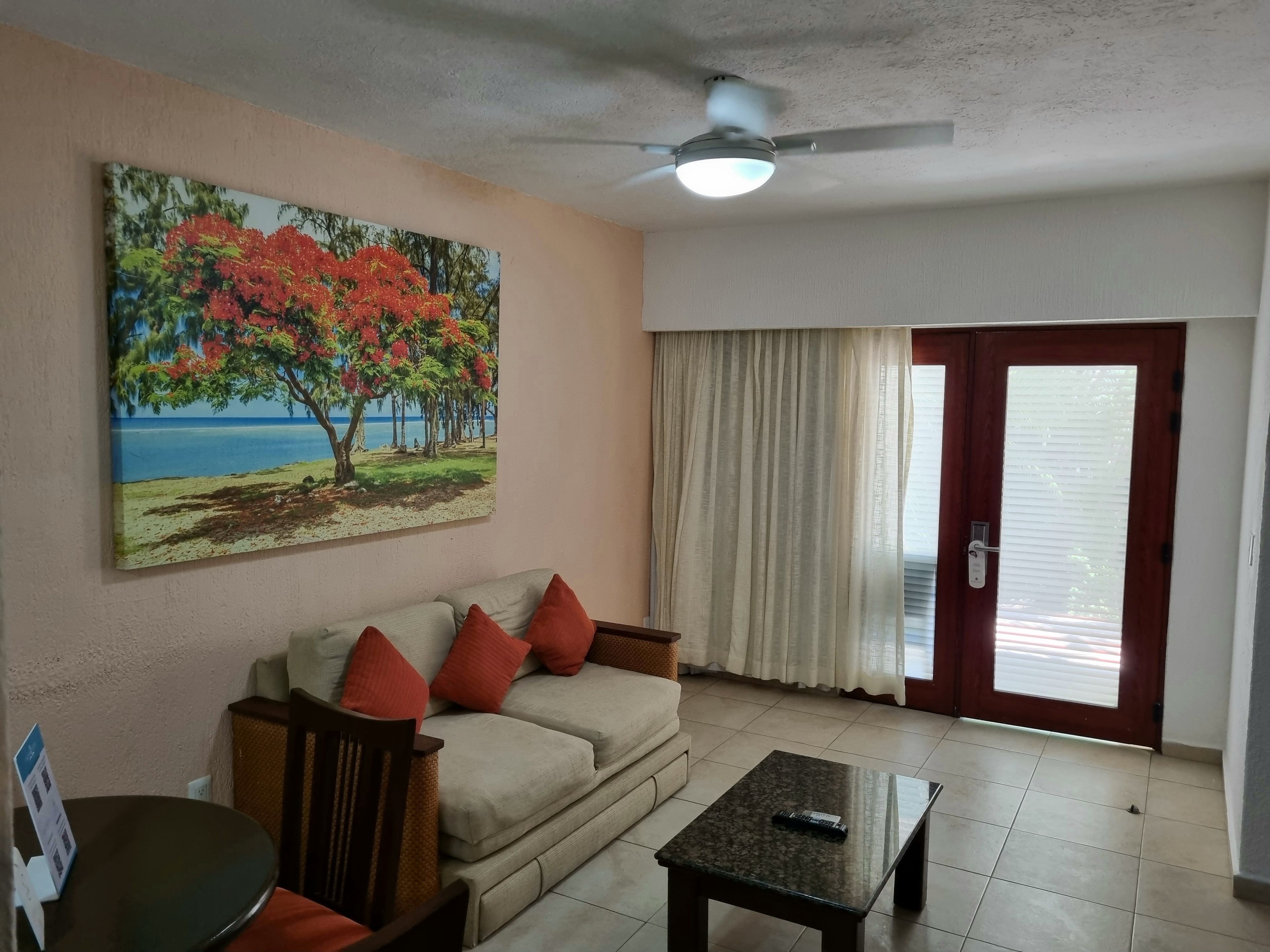 Una sala de estar con un sofá, una mesa de centro, un cuadro que muestra un árbol y una playa, un ventilador de techo y una puerta con cortinas.