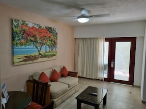 Un salon comprenant un canapé, une table basse, un tableau représentant un arbre et une plage, un ventilateur de plafond et une porte avec des rideaux.