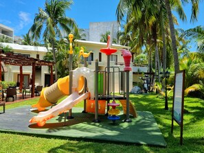 A playground with slides and climbing structures, surrounded by palm trees and a building with a balcony. - Laguna Suite (Cancun)