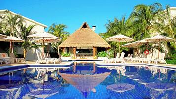 Un área de piscina con sillones de descanso y sombrillas, rodeada de palmeras y una estructura de techo de paja.