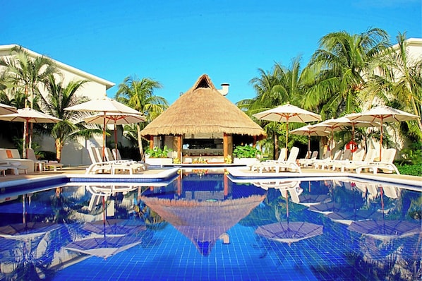 A pool area with lounge chairs and umbrellas, surrounded by palm trees and a thatched-roof structure.