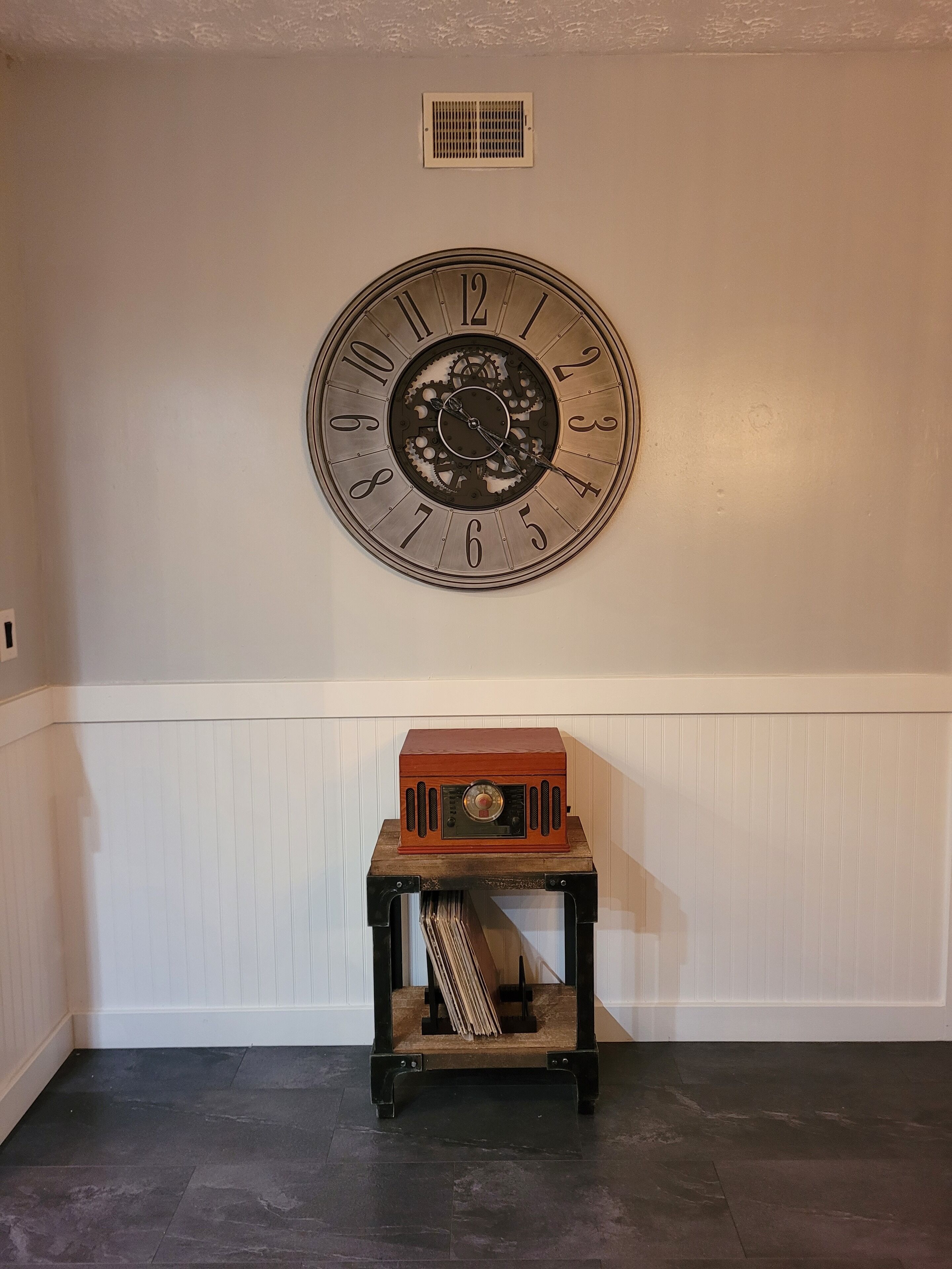 Eine an der Wand montierte Uhr im Zahnrad-Design, ein Vintage-Radio auf einem Holzständer und ein Lüftungsgitter oberhalb der Uhr.