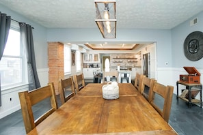 Un comedor con una mesa y sillas de madera, una cesta tejida, una pared de ladrillo y una cocina al fondo.