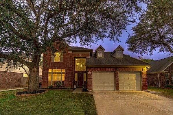 A two-story brick house with a garage, large windows, and a tree in the front yard. - Comfortable, Upscale House with a pool and spa! (Katy)