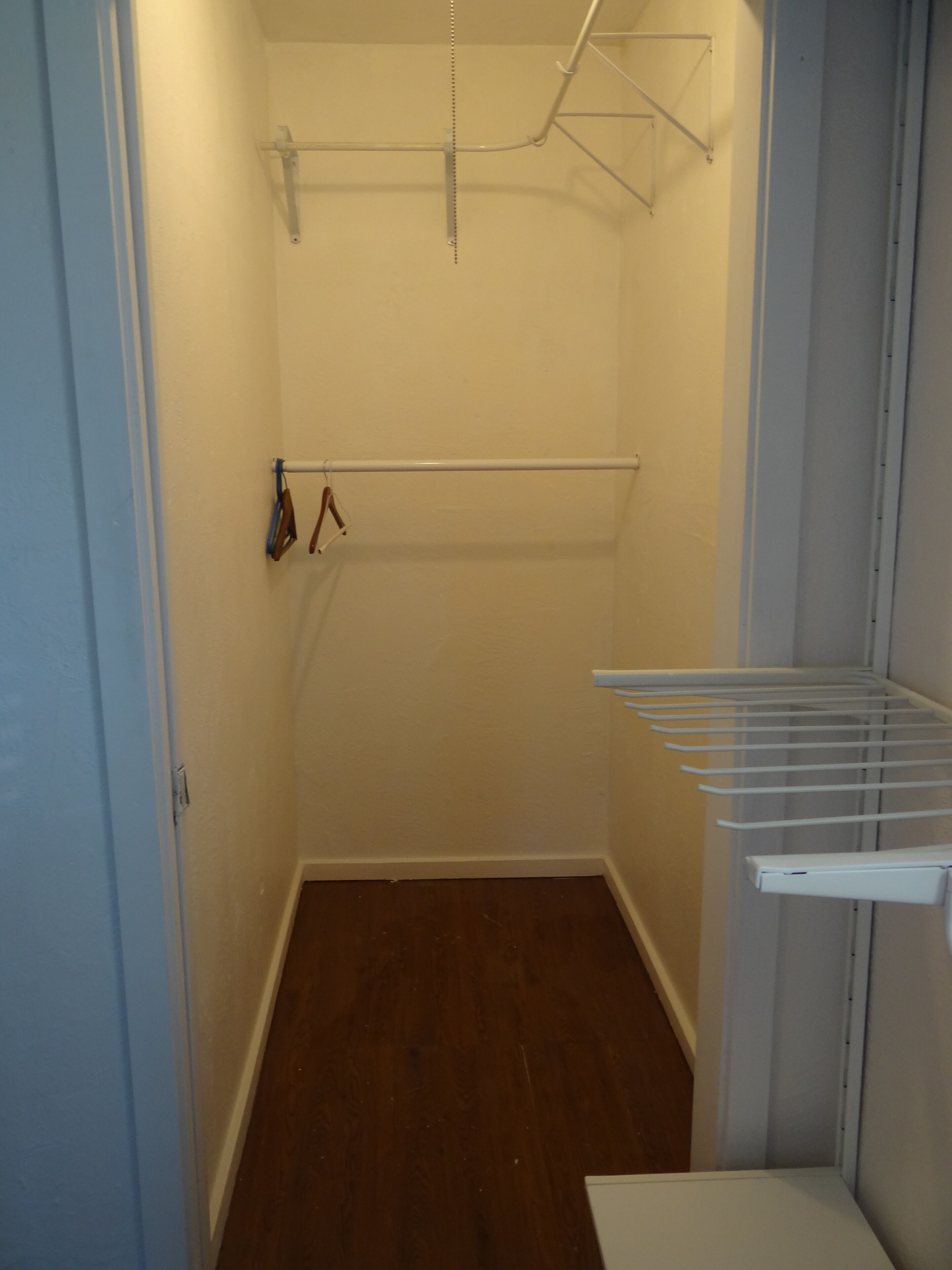 Ein kleiner, schmaler Schrank mit Holzboden und weißen Wänden. Er enthält eine Metallstange und ein weißes Regal.