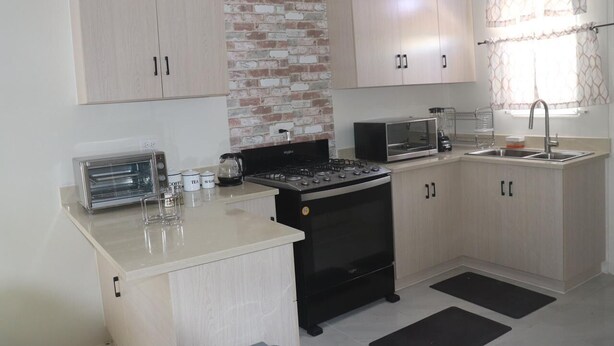 A compact kitchen with a built-in oven, a microwave, and a toaster. There are cabinets with handles, a brick accent wall, and a window with curtains.