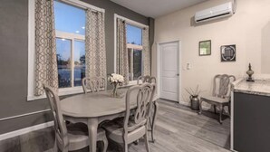 Un coin repas avec une table ronde et des chaises, de grandes fenêtres avec des rideaux, une porte blanche et un appareil de climatisation fixé au mur.