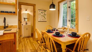 A dining area with a wooden table set for four, a window with a view of greenery, and a kitchen area with a sink and cabinets. - Hanging Rock Cabin - (Lyndhurst)