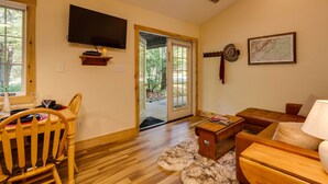 A cozy living room with a wooden floor, a television mounted on the wall, a dining area with a chair and table, and a sofa with a lamp. - Hanging Rock Cabin - (Lyndhurst)