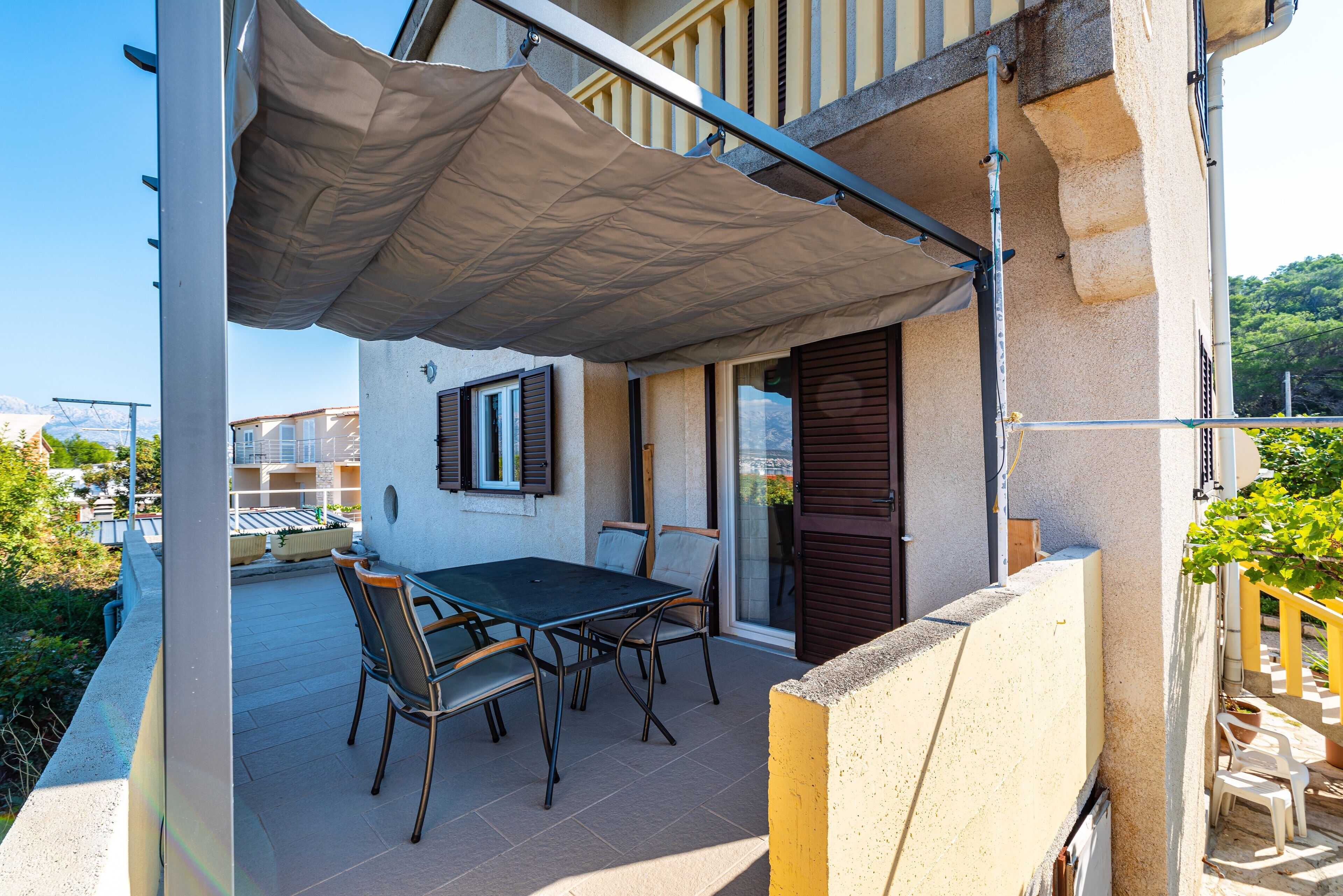 An outdoor patio with a table and chairs, a covered area, and a view of a building with windows and shutters.