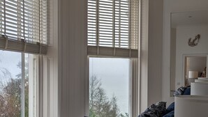 A bedroom with a large bed, a bedside table, a lamp, and a view of the outdoors through the windows. - Stylish 1 Bed Apartment with Sea views (Shanklin)