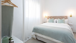 Ein ordentlich bezogenes Bett mit hellem Kopfteil und Kissen, ein Nachttisch mit Lampe und ein Kleidungsstück an einem hölzernen Kleiderbügel.