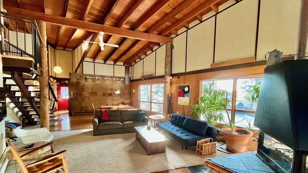 A spacious living area with a high ceiling, wooden beams, a sofa, a coffee table, a television, a fireplace, and a staircase.