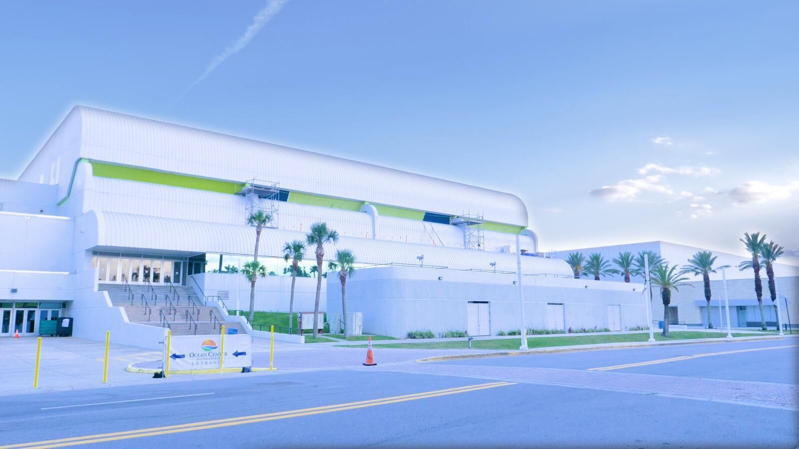 A large, modern building with a curved facade, a staircase leading to an entrance, and palm trees in the foreground.
