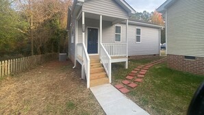 A single-story house with a front porch, a visible door, and a small front yard with a wooden fence and a paved walkway. - Budget Friendly Bungalow (Raleigh)