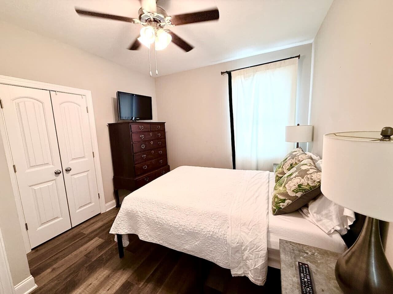 A bedroom with a bed, a dresser, a ceiling fan, a TV, and a window with curtains.