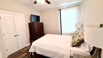A bedroom with a bed, a dresser, a ceiling fan, a TV, and a window with curtains.