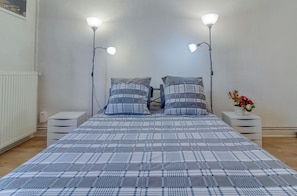 A bedroom with a plaid bedspread, two bedside lamps, a nightstand with a vase of flowers, and a radiator.
