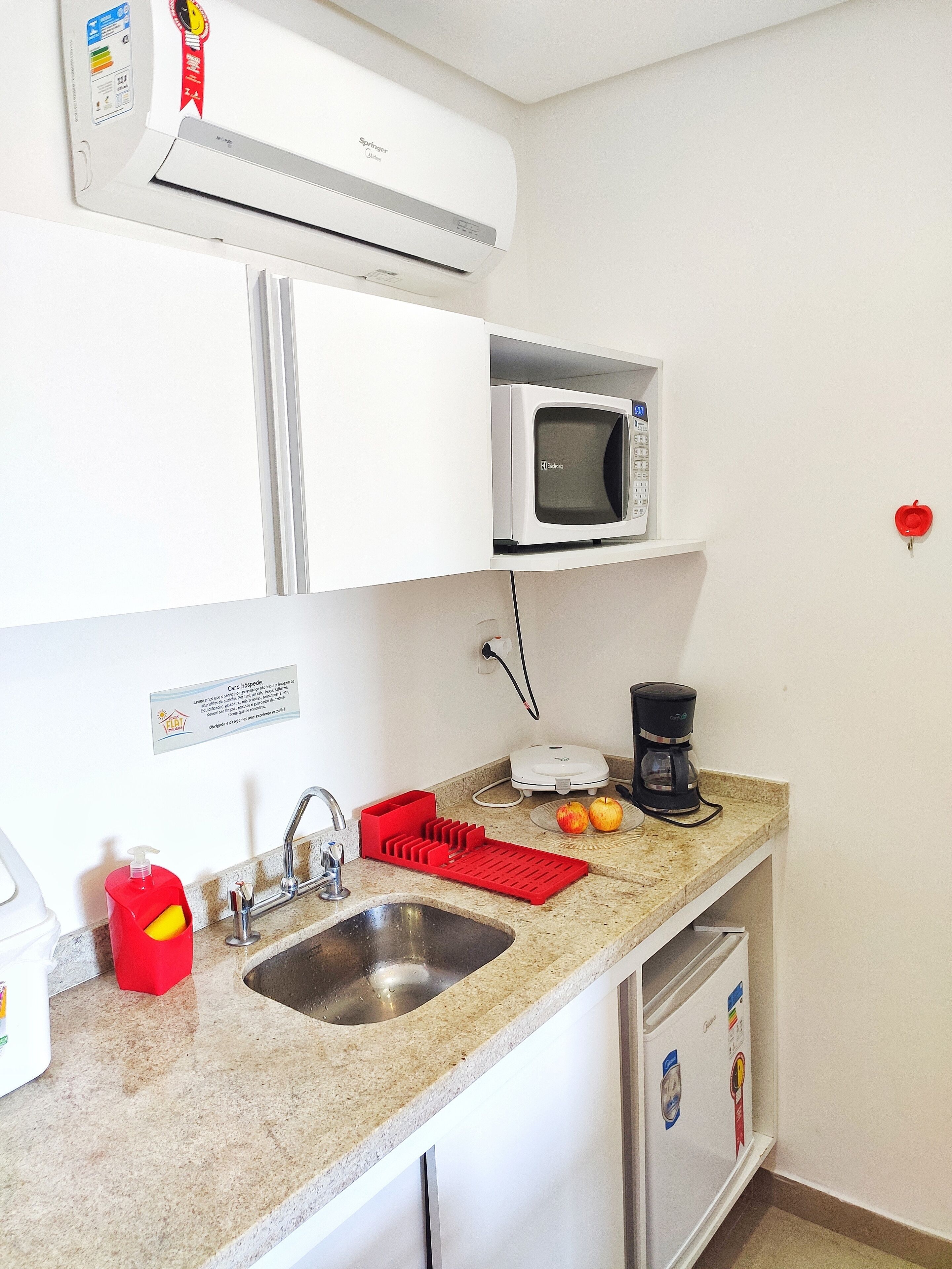 Cozinha com micro-ondas, cafeteira, dosador de sabonete vermelho e pia.