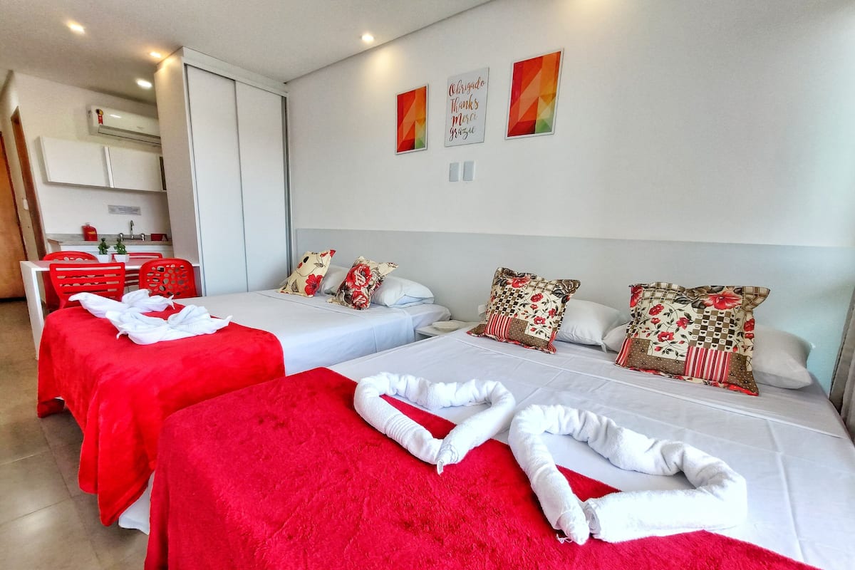 Ein Hotelzimmer mit zwei Betten, roten Handtüchern, Kissen mit Blumenmuster und einer kleinen Küche im Hintergrund.