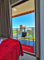 Habitación de hotel con balcón, cama con cubierta roja y vistas al mar y edificios.