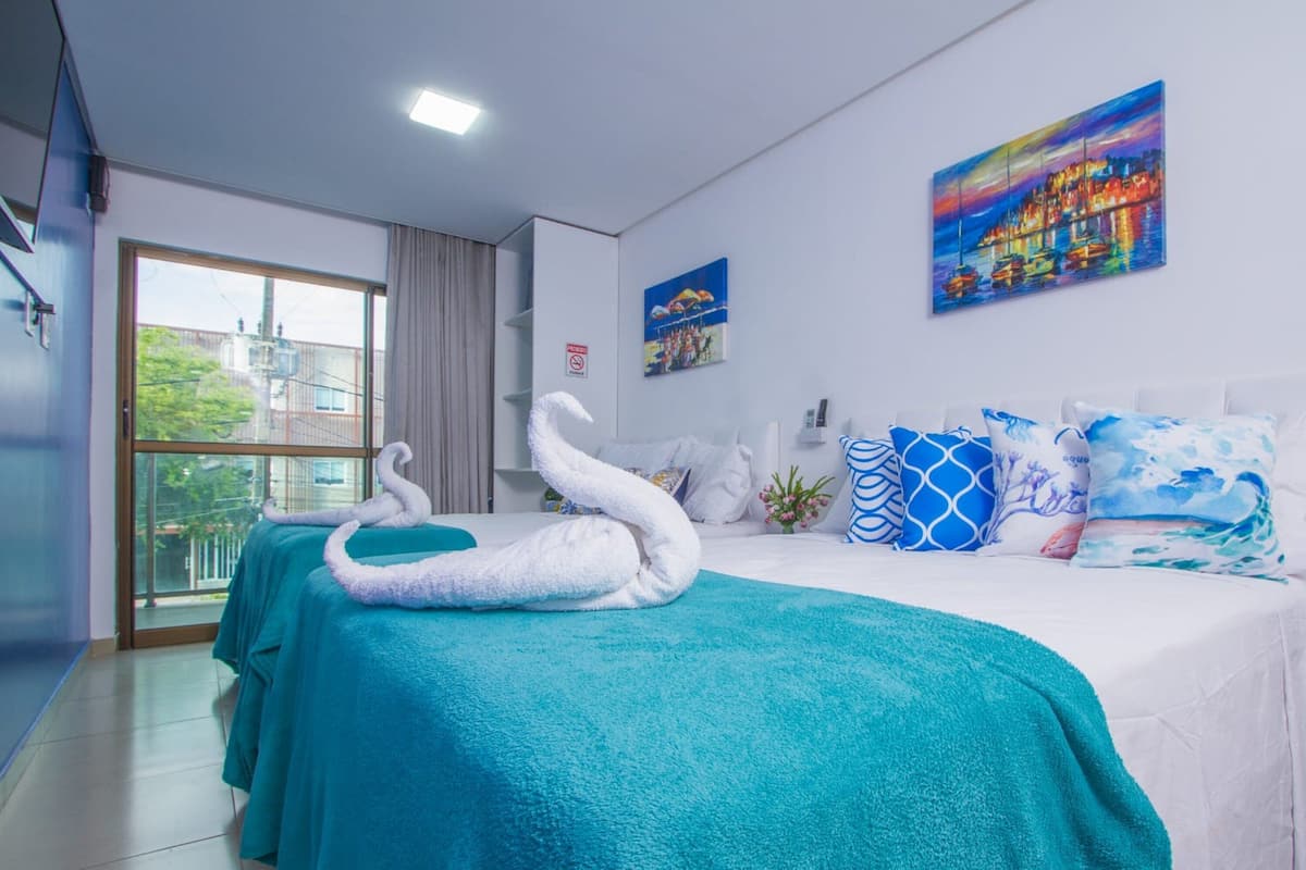 Ein Hotelzimmer mit einem Bett, zwei zu Schwänen geformten Handtüchern, petrolfarbener Bettwäsche, einem Stadtbildgemälde und Blick durch ein großes Fenster.