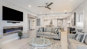 A modern living room with a flat-screen TV, a fireplace, a ceiling fan, and a dining area in the background.