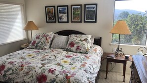 Une chambre à coucher avec une couvre-lit à motifs floraux, une tête de lit en bois, une table de chevet avec une lampe, une commode et une fenêtre donnant sur des arbres.