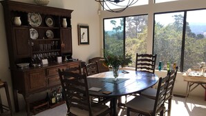 Un coin repas avec une table et des chaises en bois, un buffet contenant de la verrerie, et une vue sur les arbres à travers de grandes fenêtres.