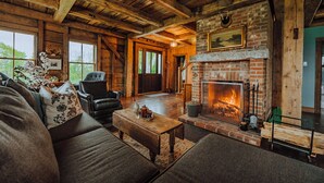 A rustic interior with a fireplace, wooden beams, leather furniture, and a view through the windows. - Kingdom Carriage House, Historic Home on 35 Acres in the Northeast Kingdom (Sutton)