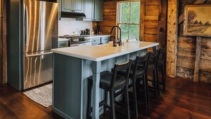 A kitchen with wooden beams, a stainless steel refrigerator, a large island with bar stools, and a window providing natural light. - Kingdom Carriage House, Historic Home on 35 Acres in the Northeast Kingdom (Sutton)