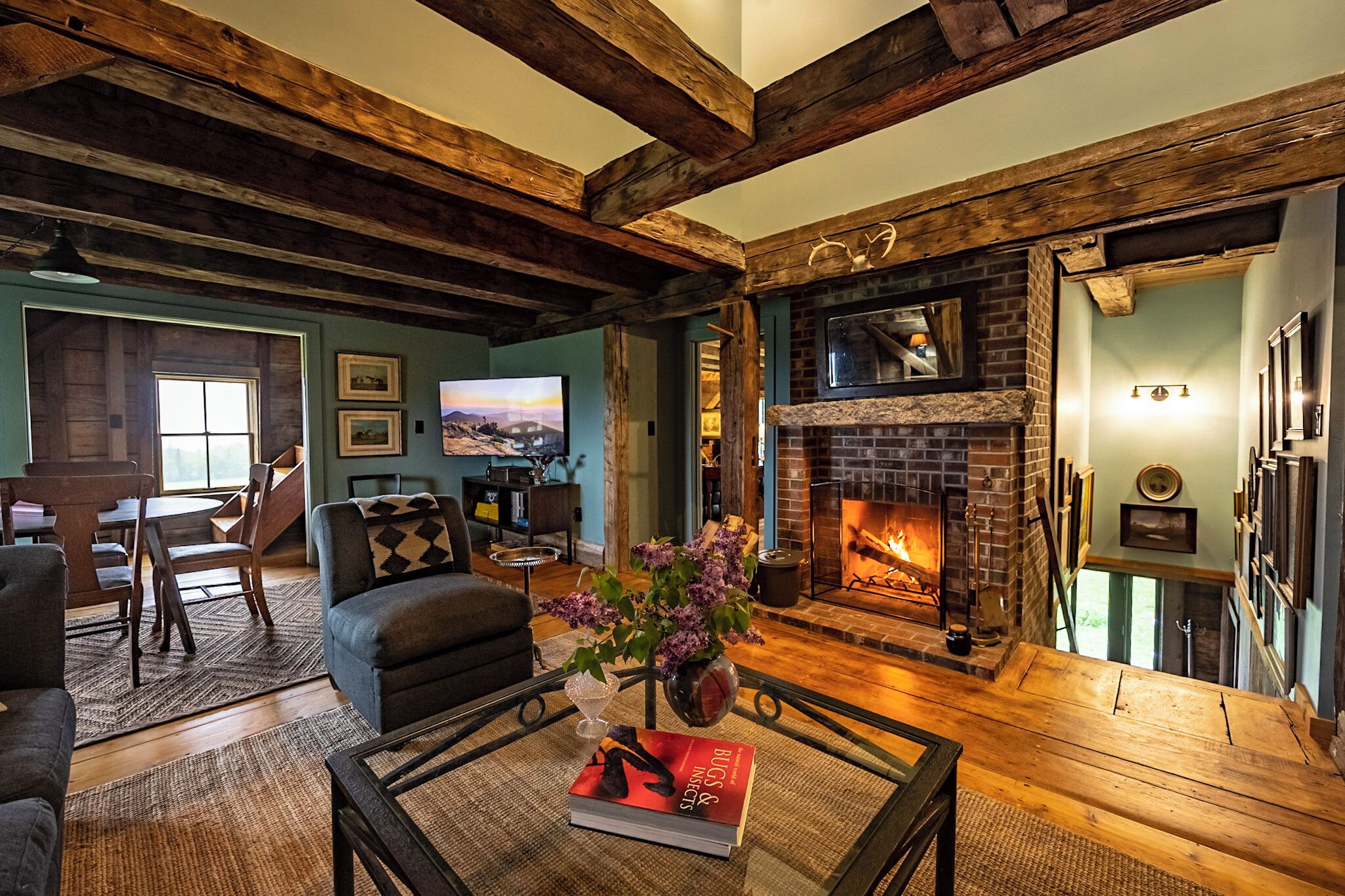 A living room with a fireplace, wooden beams, a television, and a dining area with a table and chairs.