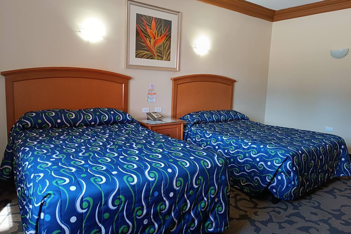 Habitación de hotel con dos camas, cada una con cubrecama de estampado azul y verde, cabecero de madera, mesita de noche con teléfono y un cuadro enmarcado en la pared.
