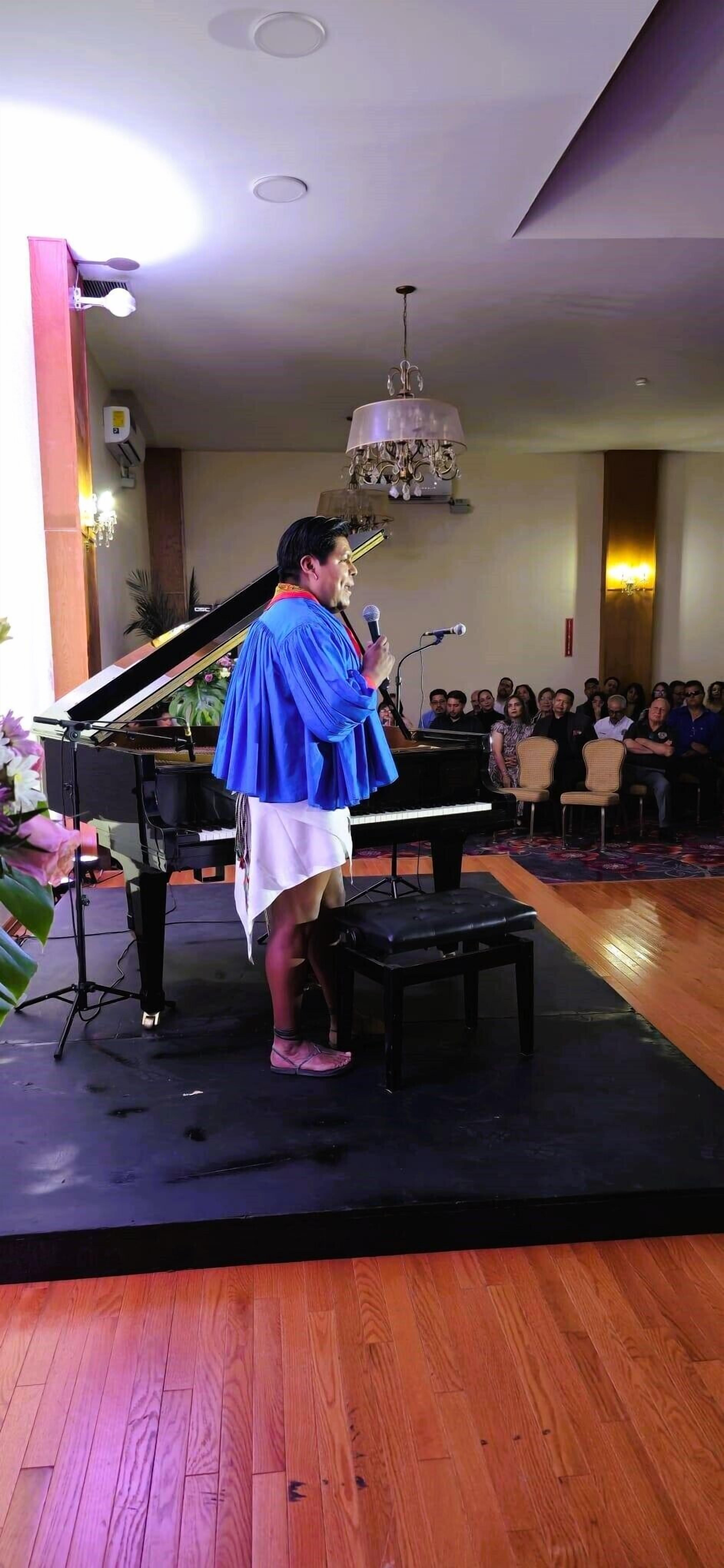 A person standing on a stage with a piano, holding a microphone, in a room with an audience seated.